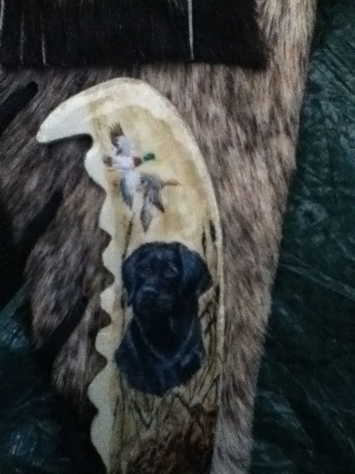 The handle of a knife shaped and painted to be like animal teeth. Painted on the handle is a black lab standing in cattails and long grass as two ducks fly overhead. The knife handle is laid on an animal pelt.