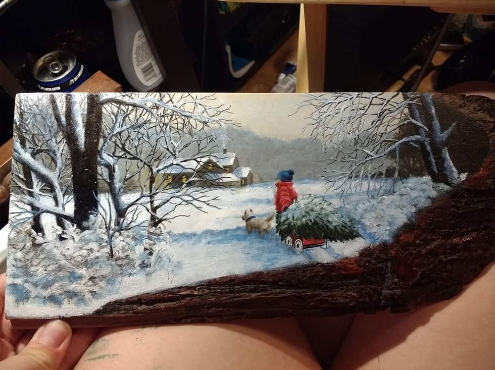 A painting on a slice of wood with some of the bark still on it, laid across someone's lap. The painting shows a snowy winter scene with a child and a dog. The child is pulling a wagon laden with a christmas tree, and both are headed towards a warm looking house in the distance with smoke rising from the chimney and lights in the windows. Freshly fallen snow blankets trees and bushes around the child. 