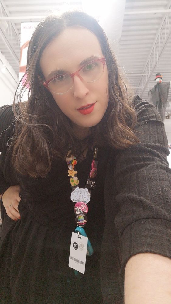 Trans woman wearing a black dress with a black throw. She has bright red lips that match her red glasses. From her neck dangles a lanyard covered with enamel pins. The stickers on her badge say "Try Me"