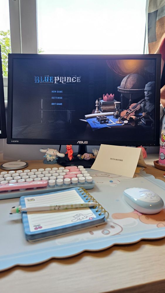 A picture of a desk with a notebook on it open with the title screen for video games Blue Prince on the monitor. 
