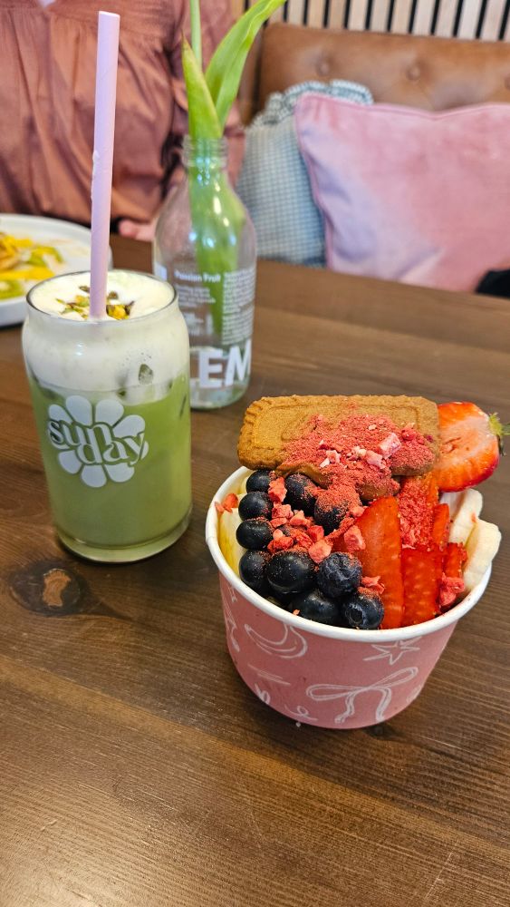 A picture of a table with an iced matcha drink and a pink bowl of fruit with acai smoothie underneath. With a lotus biscoff biscuit on top. 