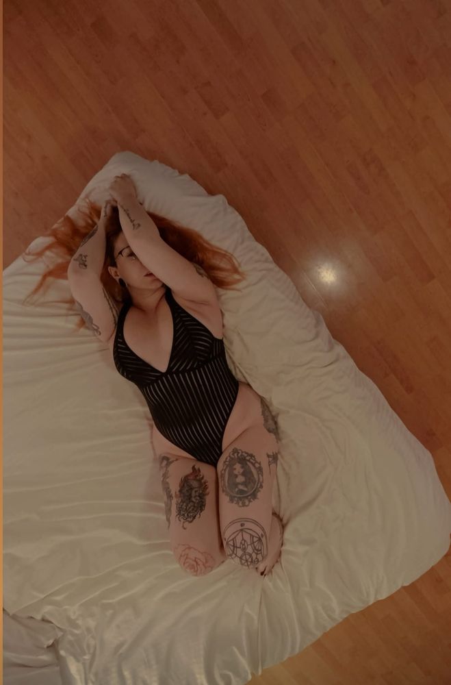 Ginger-haired Rena wearing a black bodysuit lying down sensually in a bed with her arms up