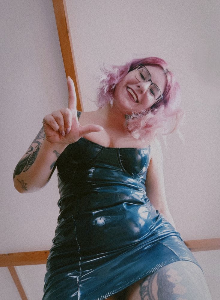Mistress Rena looking down at the camera with a smile and making the L symbol with her hand