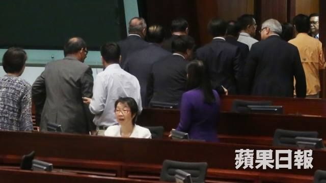 A bespectacled woman (pro-Beijing trade unionist and lawmaker Chan Yuen-han) sits with her mouth agape in the chamber of Hong Kong's Legislative Council while her colleagues head out of the chamber through a door behind her.
