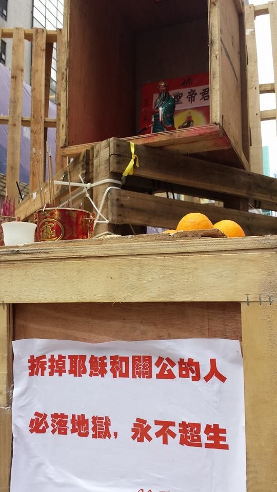 A makeshift wooden shrine to the Kuan Ti with a ceramic statuette of the deity, incense and some oranges presented as offerings in front of it. Below is a sign that reads in Chinese: Whoever removes Kuan Ti and Jesus will be condemned to hell, never to be reborn.