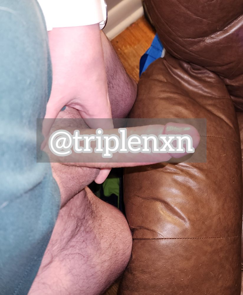Triple standing over a brown leather chair, text overtop of his cock, his legs pressing slightly against the chair and his hand holding his cock and balls in place 