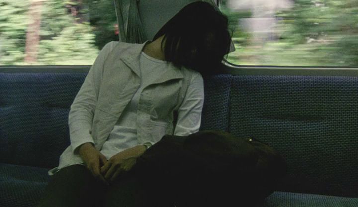 Still from Hirokazu Kore-eda’s film Distance. It shows a woman asleep on a train. She is wearing a white long sleeve shirt, cream colored vest, and dark pants. She is slumped in the seat and her hair is covering her face. 