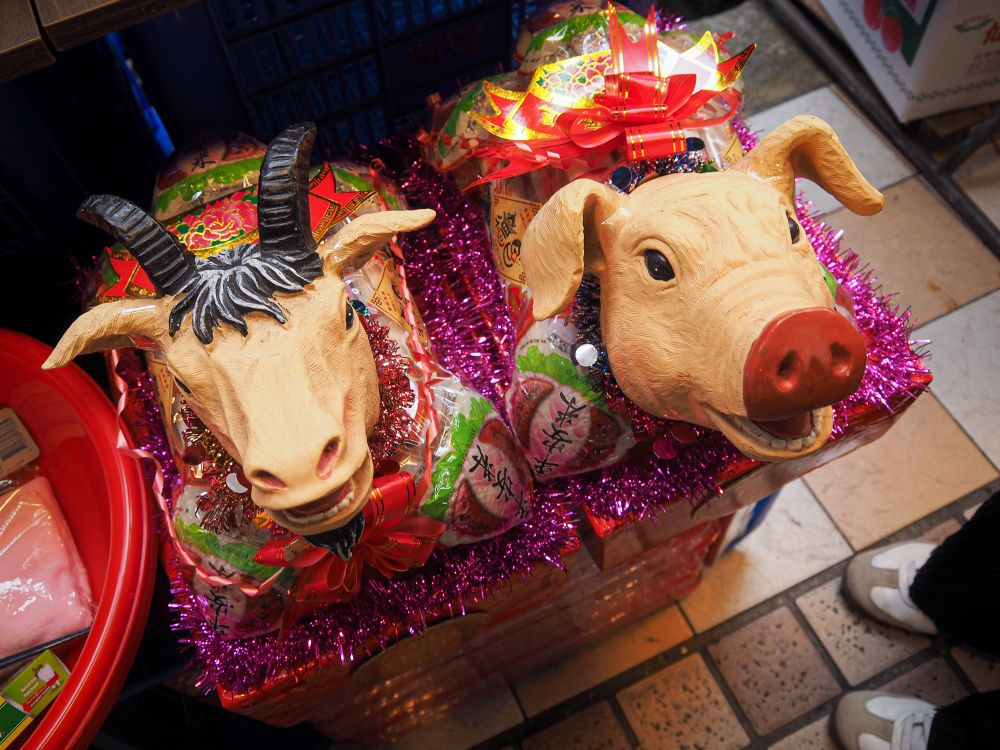 A colorful display of ceremonial offerings at a market stall, featuring plastic animal masks including a cow with black and white markings and a pig with a red snout, surrounded by shimmery purple tinsel garland, red ribbons, wrapped candies and snacks, and traditional paper decorations with Chinese characters.