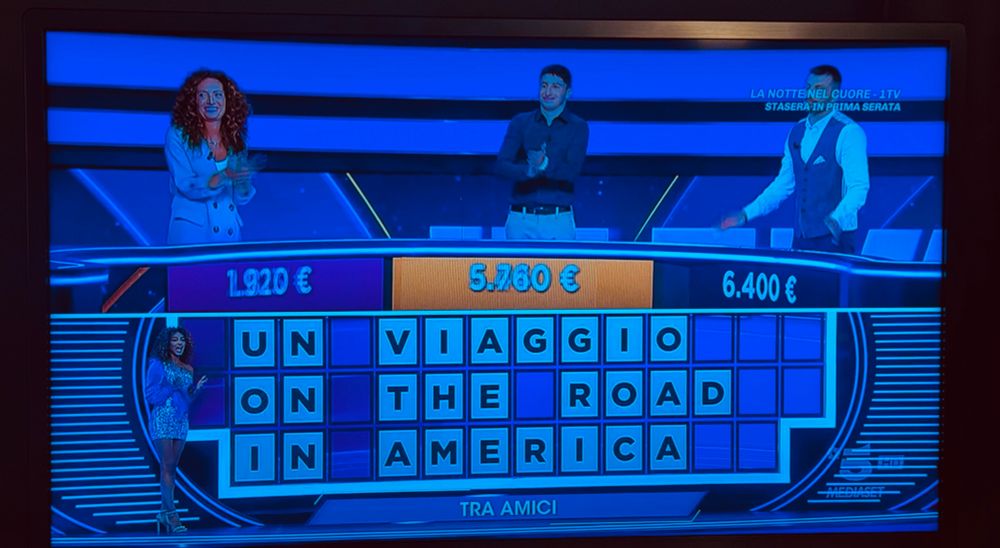 Pic of the game show. There normal or perhaps giant people on the top at podiums. One very tiny woman by a board of letters below 