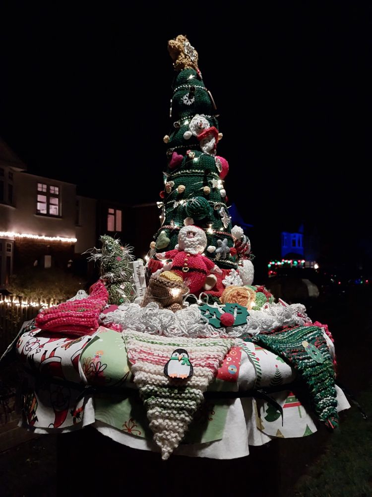 A knitted post-box topper in the dark, shaped like a Christmas tree with figures of Santa and other things, surrounded by knitted bunting.