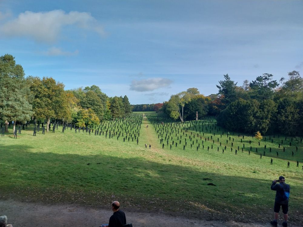 Green field sloping away from the viewer with black silhouette figures of service men, some people in foreground viewing the installation. Blue sky, trees in background, path mown between the figures with viewers walking along.