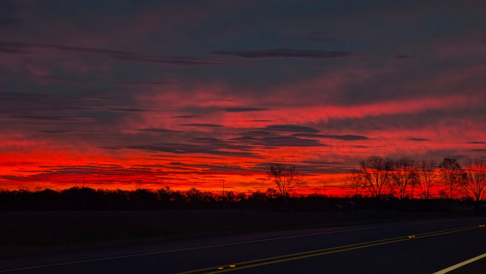 photo of pre-dawn light in the sky. clouds are a red and trees are shown in silhouette. in the foreground you can see the surface of the highway 