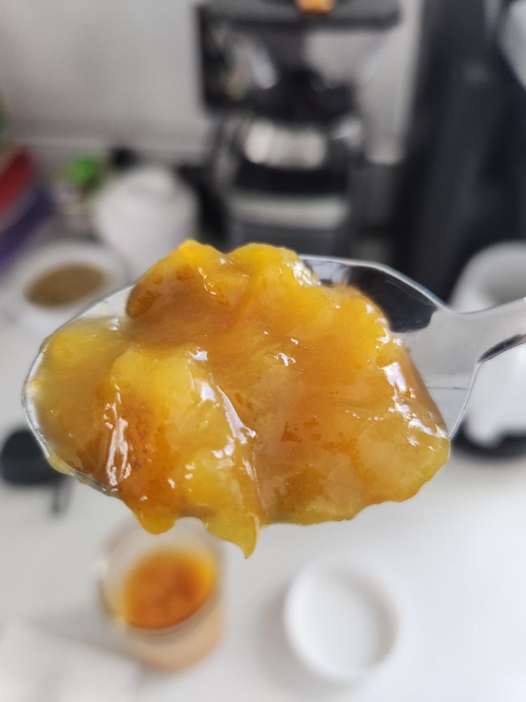 A spoon with freshly made calamondin marmalade in front of a white countertop with blurred objects in the background.