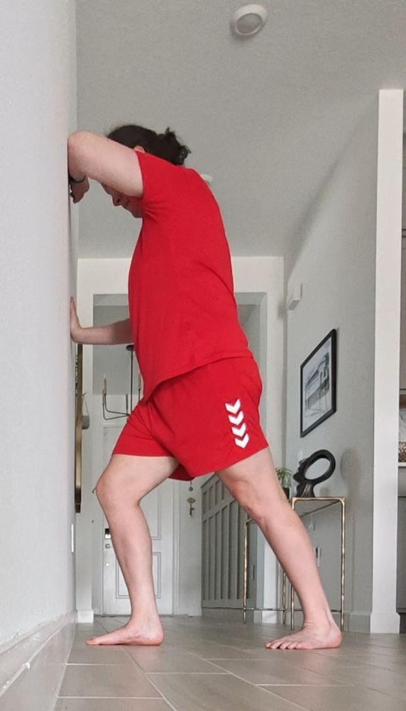 A lady in red sports uniform with her arms against a white wall. Her right foot is close to the wall and her left leg is back and straight.