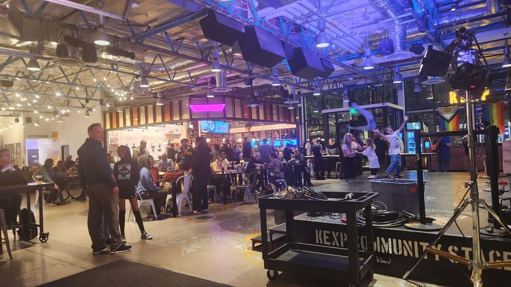 The KEXP Gathering Space, a large room with people seated at tables. In the back corner is Cafe Vita, a coffee shop, and in the front is a stage. It is dark outside. 