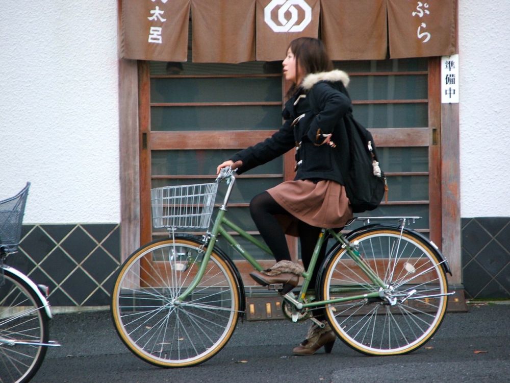 A woman with a backpack sits astride a mamachari, which is a Japanese bicycle with a low top bar, cute basket in the front and storage rack on the back. The woman is wearing fun shoes.