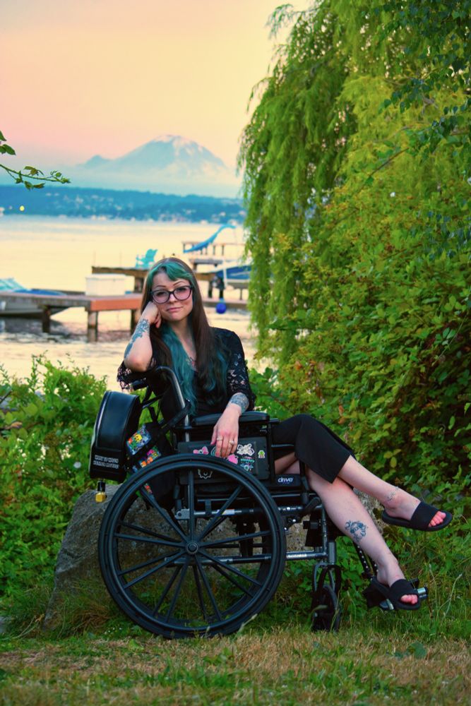 Vixey Vex in a black dress sitting in a wheelchair in front of a lake at golden hour with Mt. Rainier in the background