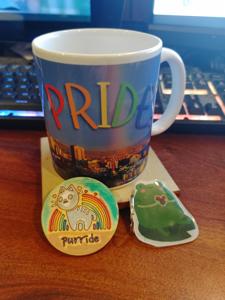 A mug with a photo of rainbows over the Seattle skyline, a wooden button with a picture of a cat and a rainbow that says 'purride,' and a shrinky-dink keychain of a bisexual frog