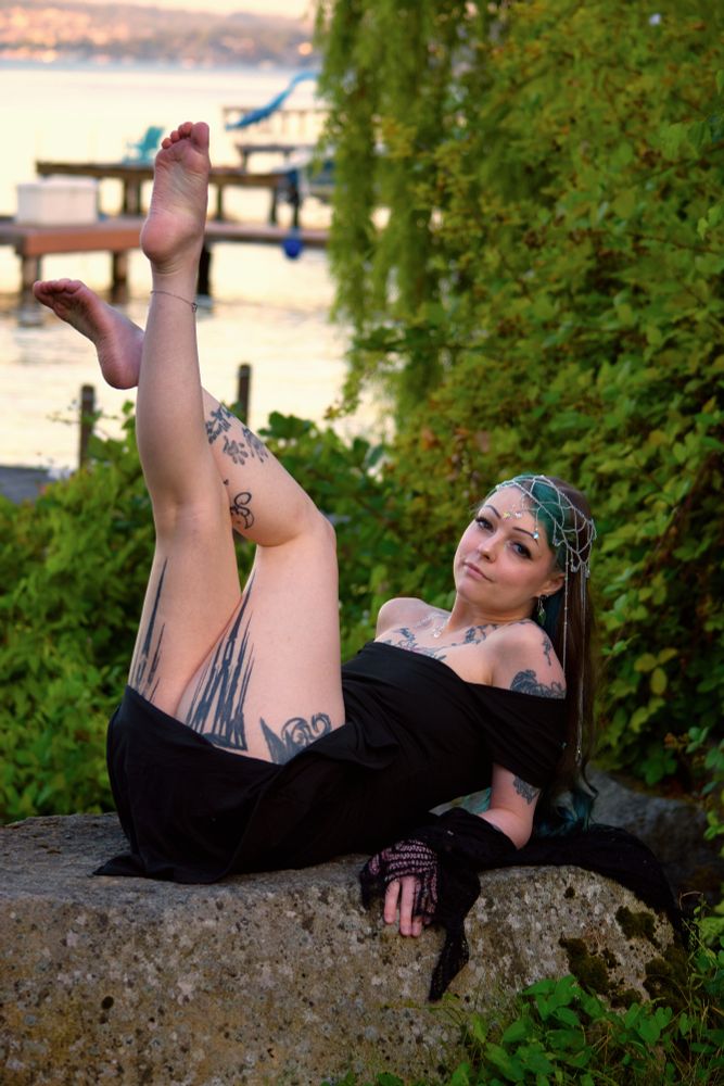 Vixey Vex in a black dress laying back on a large rock in front of a lake at golden hour