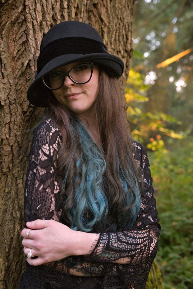 Vixey Vex in a black lace top and black hat leaning against a tree in the forest