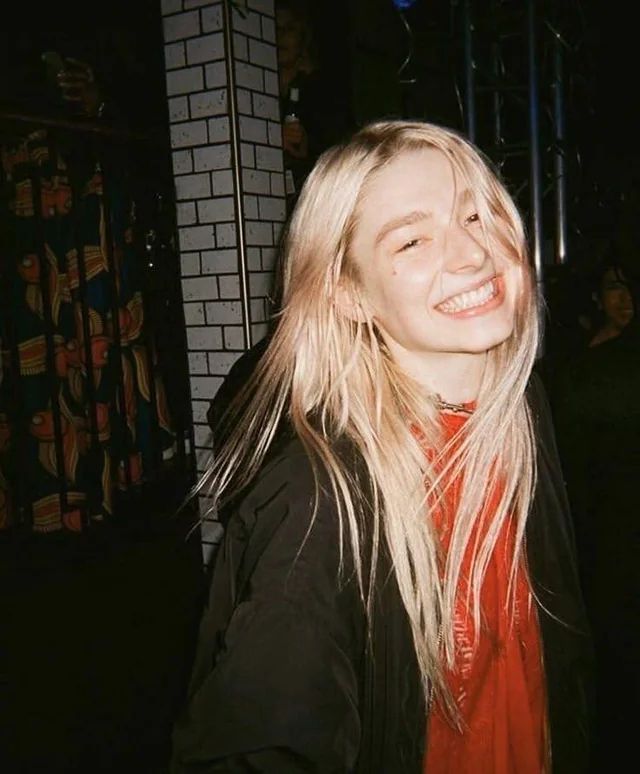 hunter Schafer, long blonde hair ,looking at the camera and smiling with teeth, wearing a red undershirt and a black jacket.