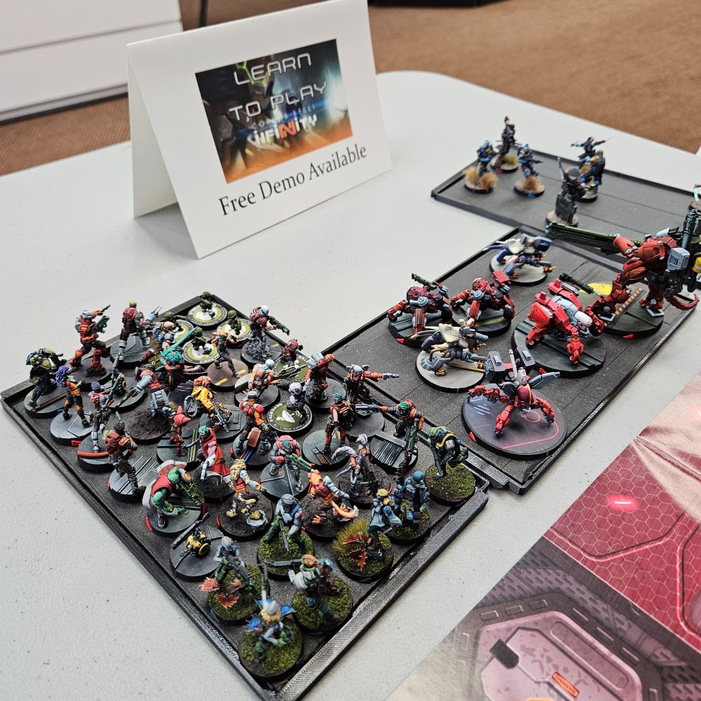 A picture of a table setup for a demo of the miniature wargame Infinity. The image is focused on a selection of painted miniatures on black trays and a paper tent advertising that this is a free learn to play demo.