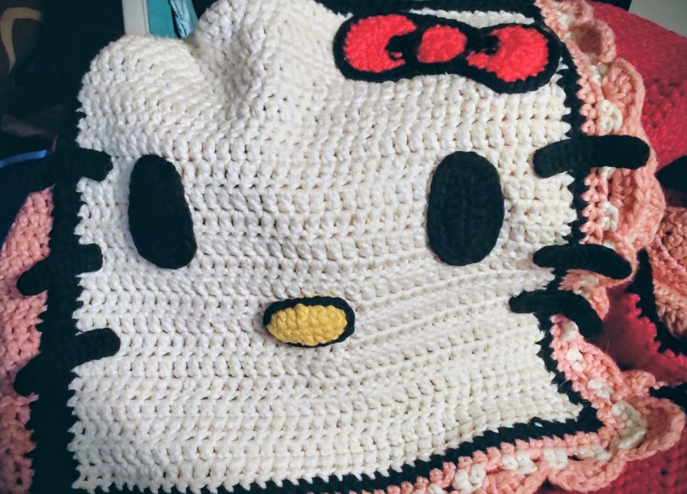 photo of a crocheted blanket, one square of which is made into a Hello Kitty. she has black eyes, black whiskers, a yellow and black nose, and the quintessential red and black bow. you can see a black, pink, and white border on the edge, and hints of pink and magenta squares on the rest of the blanket.