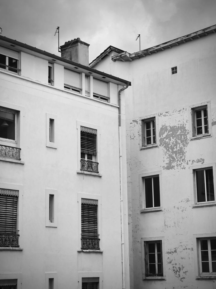 Vue (bouchée) de fenêtres sur cours. On voit 2 immeubles en angle. Et tout en haut : un petit bout de ciel.
Photo en noir et blanc