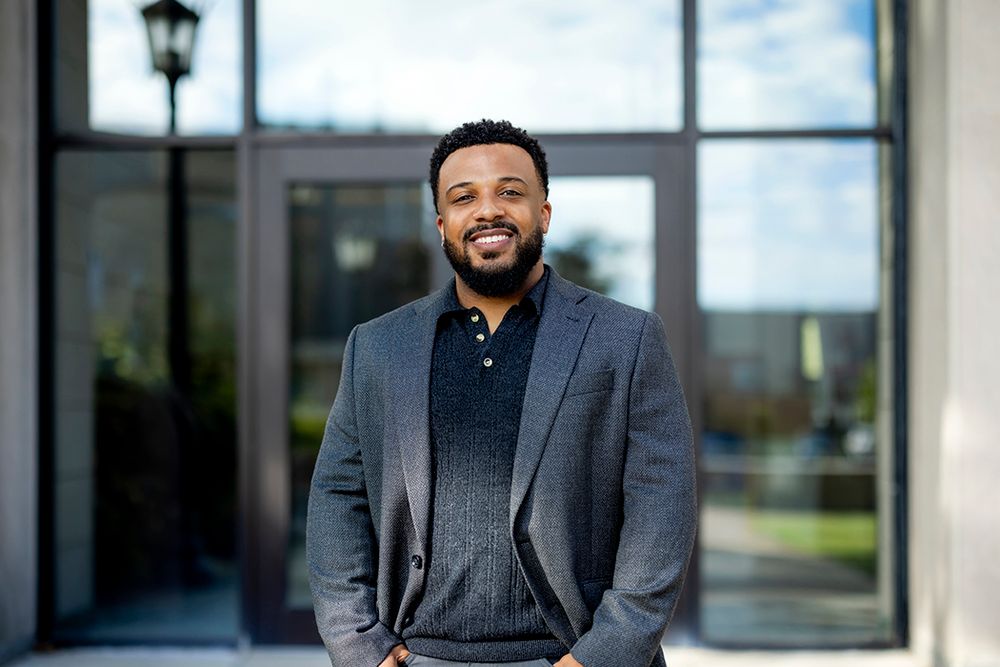 Ed-Dee G. Williams wearing a dark gray suit and black shirt smiling at the camera standing in front of a building with his hands in his pockets 