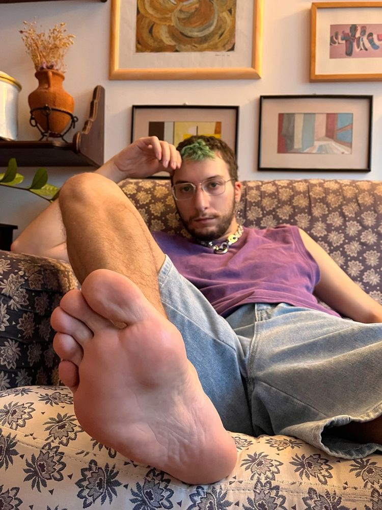 I’m lounging on the couch in a purple tank top and denim shorts, one leg lifted with the sole of my bare foot close to the camera.