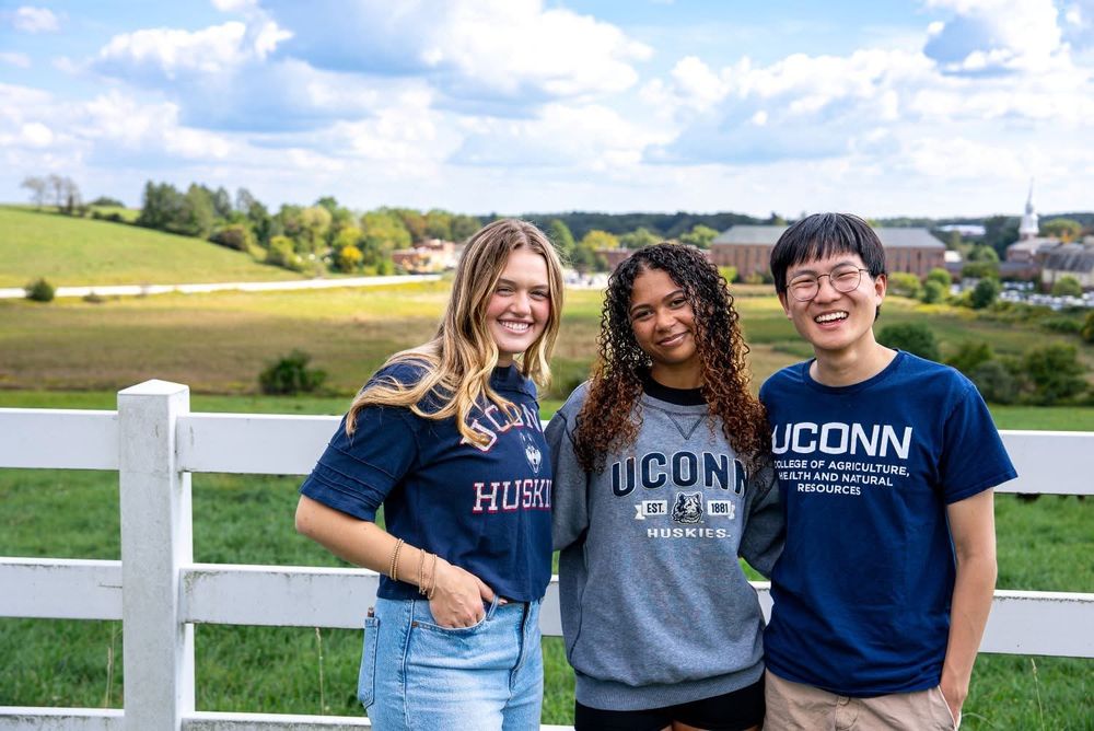Three students stand together smiling in front of a white fence overlooking a green pasture on a sunny day. All three wear UConn-branded shirts, and campus buildings and a blue sky with clouds are visible in the background.