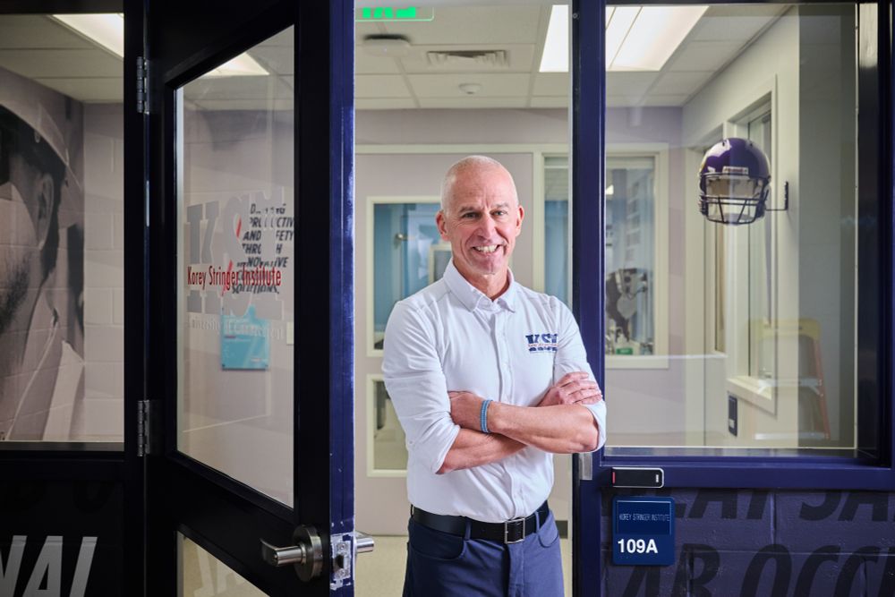 A man stands smiling with arms crossed in the doorway of the Korey Stringer Institute at UConn, wearing a white UConn KSI shirt.
