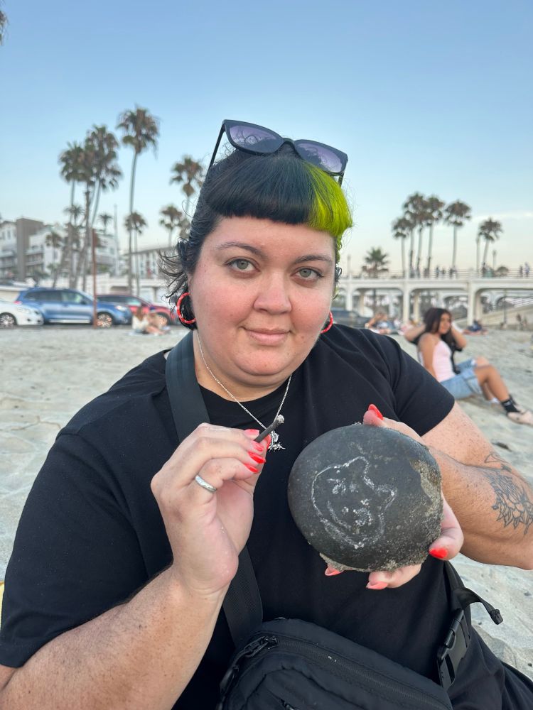 Katie holding a black rock with a cat face drawn in white pigment 