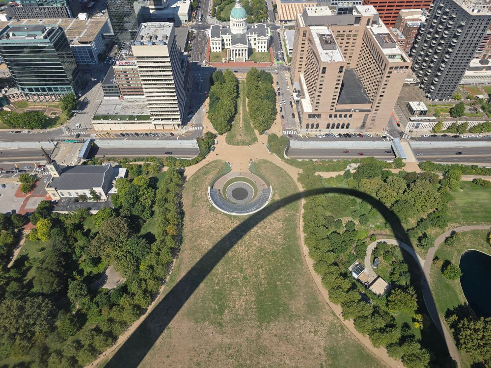 Photo taken through the window on the north leg and west side of the Arch in Gateway National Park in St. Louis, MO. The circular impression of the entrance can be seen at center with the shadow of the Arch itself just touching the edge at about 5 o'clock and moving off the bottom of the photo. There is a green grassy strip leading to a big white building at the top of the photo. This is, I think, the Old Courthouse building. The top half of the photo shows city buildings and streets, while the rest of the bottom half is trees, grass, and walking paths.