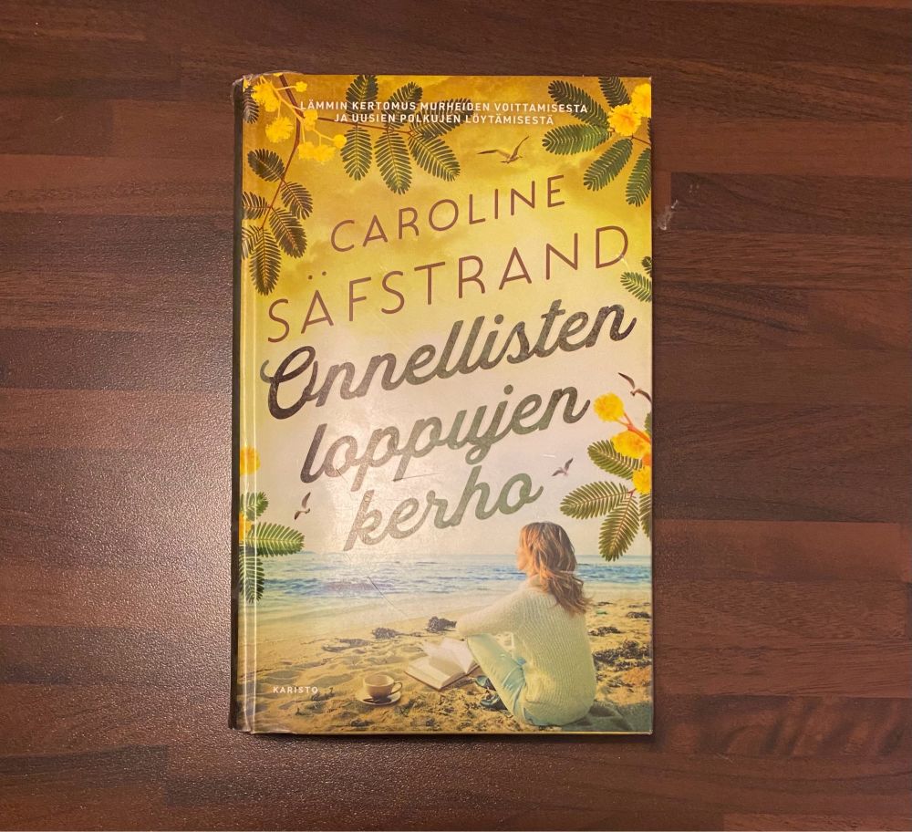 Kansi kirjasta Onnellisten loppujen kerho. Kirjailija Caroline Säfstrand. Kannessa nainen istuu yksin hiekkarannalla vaaleissa vaatteissa edessään kirja ja suuri kahvikuppi.
