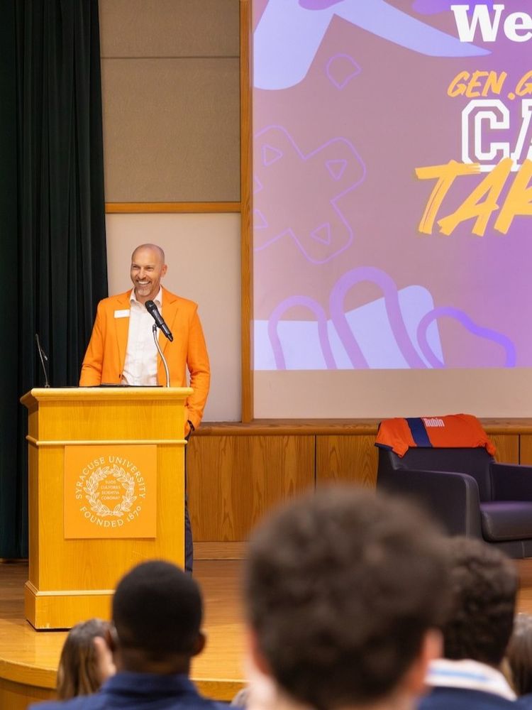 Person speaking at a podium with a Syracuse University logo, addressing an audience in a lecture hall, with a large screen displaying text "Welcome Gen. G" in the background.