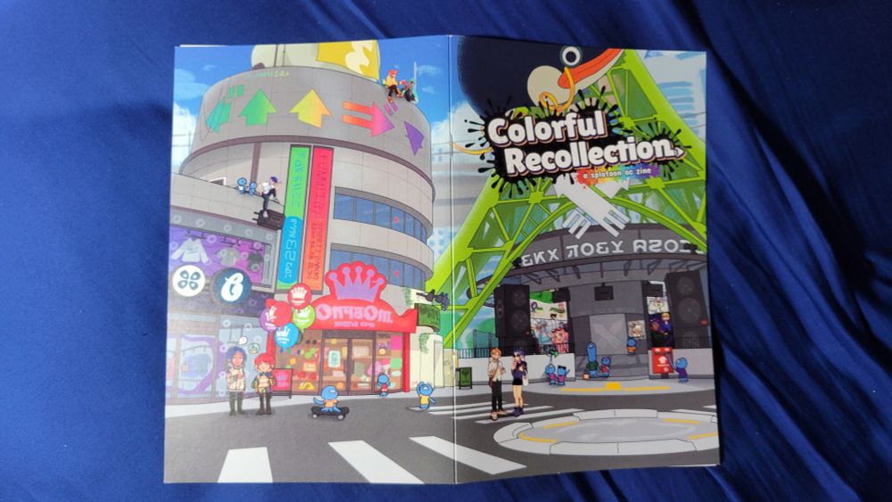 a photograph of the front and back cover of my friends and i's splatoon oc zine, colorful recollection. the cover art depicts inkopolis plaza, with our ocs and jellyfish scattered in different locations.