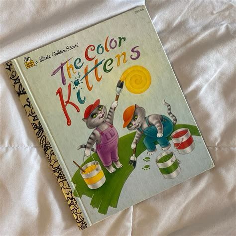 A Little Golden Book on a white cloth.  The book is titled The Color Kittens and shows two gray cats - one in purple overalls and one in blue overalls painting.  One is painting the yellow sun and the other the green grass 