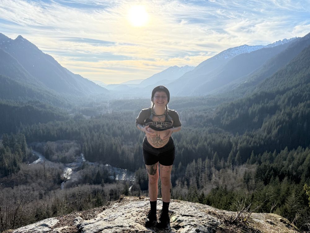 Faye stands in front of a panoramic view on a cliff ledge, tits out and grinning