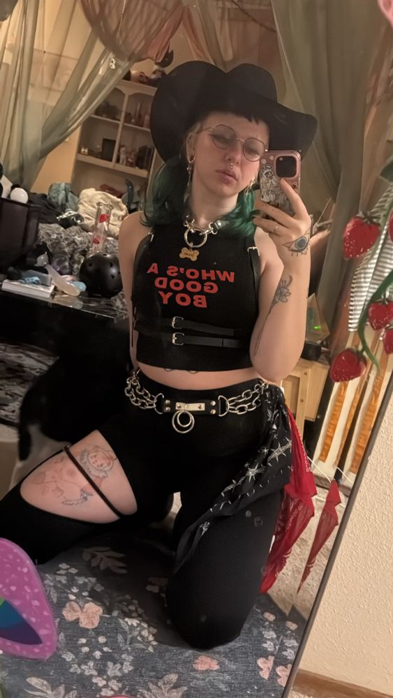 Faye in a black cowboy hat, hair in pigtails, crop top that says “who’s a good boy”, chest harness, strappy pants, chain belt flagging red and black on the right, doc martens. Posing for a mirror selfie. Kneeling. 