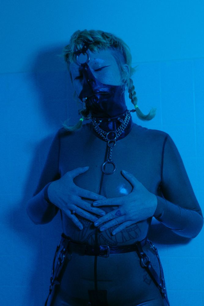 Faye stands in a shower wearing a translucent black catsuit and a latex mask. Their hands are covering their chest. Blue lighting. 