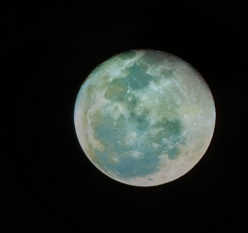 A high-resolution photograph of the full Moon against a pitch-black sky, captured with enhanced colors revealing subtle mineral variations—tones of teal, green, and gold swirl across its cratered surface.