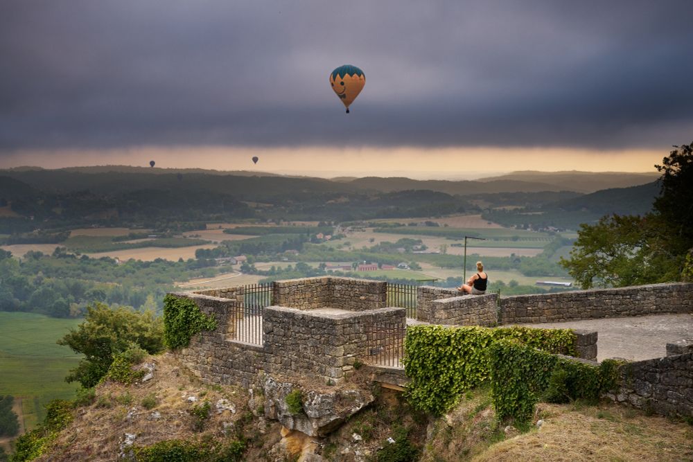 A person sits on the stone walls of an old cliffside fort, overlooking a vast valley at sunset. Several hot air balloons drift through the dramatic sky, with one large colorful balloon standing out against dark clouds and golden light.