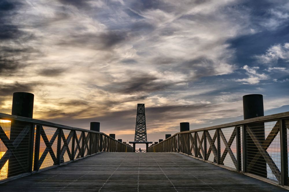 A wooden pier with repeating X-pattern railings and evenly spaced vertical posts leads to a sculptural tower under a softly colorful sunset sky. The composition emphasizes symmetry and alternating design elements.