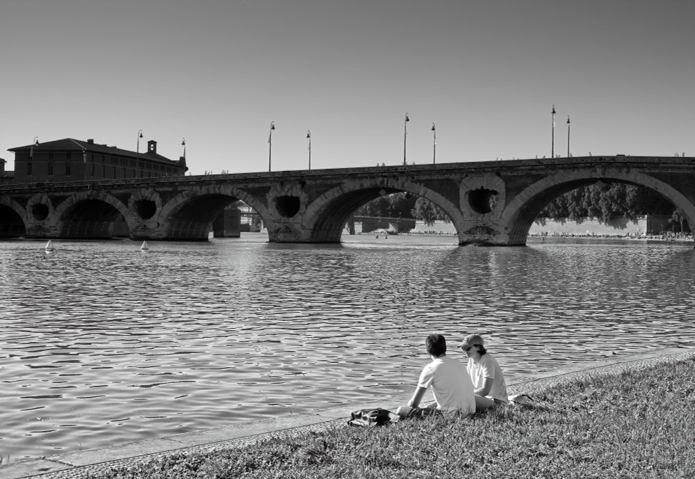 A black-and-white photograph of a historic stone bridge spanning a calm river. In the foreground, a couple sits on the grassy bank, facing the water. The bridge's arches reflect gently in the rippling surface, while soft light casts long shadows, creating a tranquil and timeless atmosphere.