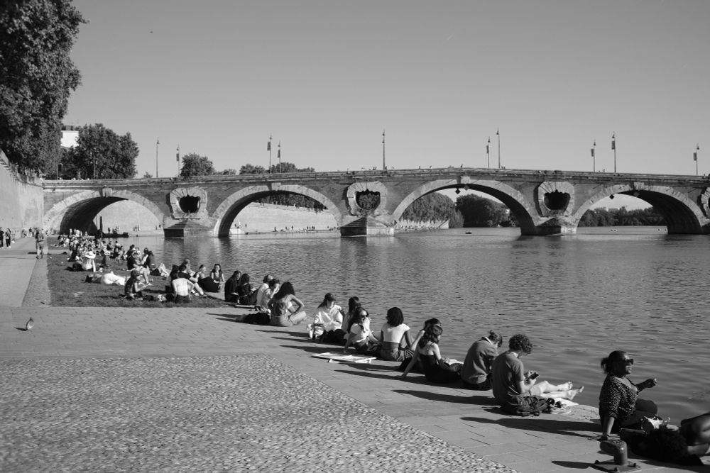 A black-and-white photo of people sitting along a riverbank near an arched stone bridge. The scene shows alternating groups and individuals, with repeating bridge arches and light-shadow contrasts throughout.