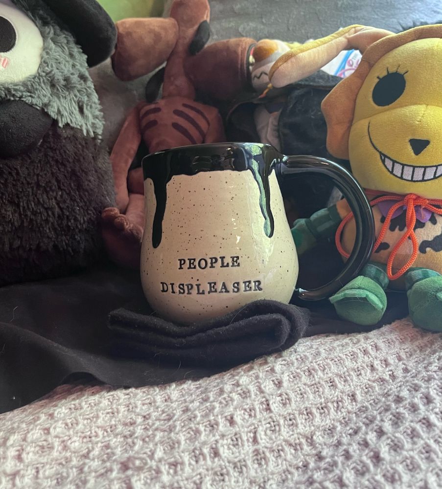 Nestled in between plushies, a homemade mug sits within them all with “People displeaser” stamped on the front, with black glaze oozing out of the top.