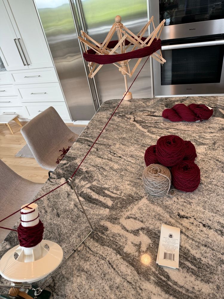 A yarn winding setup is spread out across a kitchen island with a granite countertop. A burgundy skein is stretched on a wooden swift, feeding yarn to a white and green ball winder that’s mid-spin. Several completed yarn cakes in burgundy and one in a taupe-gray shade are stacked nearby, along with an empty yarn label. Two gray chairs and a modern kitchen with stainless steel appliances are visible in the background. The scene suggests preparation for a long weekend of knitting. #knitting #knittersofbluesky #knitsky #fiberart #yarn #wool #cascade220 #aran #knit 