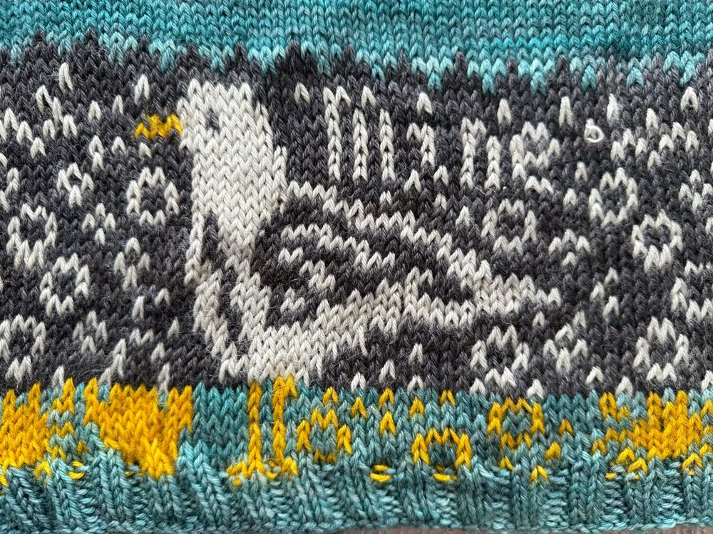 A close-up of the lower hem of the Mine sweater, showing detailed colorwork in Madelinetosh DK yarn. The design features a puffin motif in cream, black, and gray, with a bright yellow beak. Behind the puffin, the word “Mine” is subtly worked into the dark background. Below the puffin, a band of teal ribbing anchors a cheerful border of yellow flowers and teal stems, adding a vibrant contrast to the grayscale pattern above. The stitches are even and crisp, highlighting the intricate intarsia and stranded knitting techniques used.
