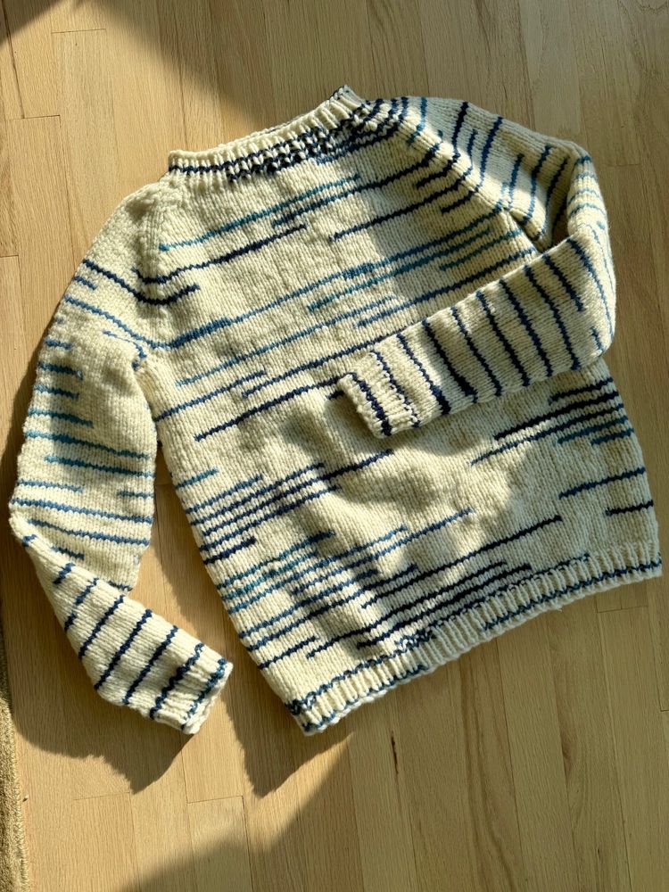 A hand-knit Galatee sweater lies flat on a wooden floor, catching the warm afternoon light. The sweater is made with Berroco Dash yarn, a cream-colored base with intermittent navy, denim, and sky-blue speckles and stripes that create a painterly, abstract look. Knit this weekend during an unexpected internet outage, the project was powered by three audiobooks and a whole lot of uninterrupted crafting time. #BerrocoDash #GalateeSweater #KnittingWeekend #AudioBookAndKnit #KnittersGonnaKnit #SlowFashion #HandmadeWardrobe #UnplugAndCreate #yarn #wool #craftiverse #craftsky #knitsky #knit #knitting #showmeyourknits #knitters #Berroco #sweater #weekend #stressknitting #travelprep